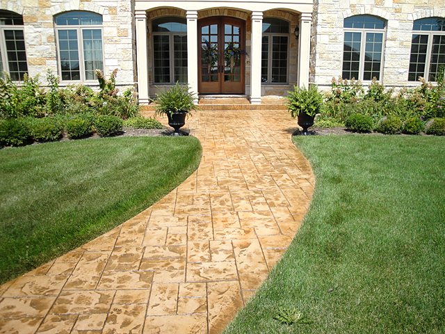 Curved tan stamped concrete walkway leading to a stone home entrance